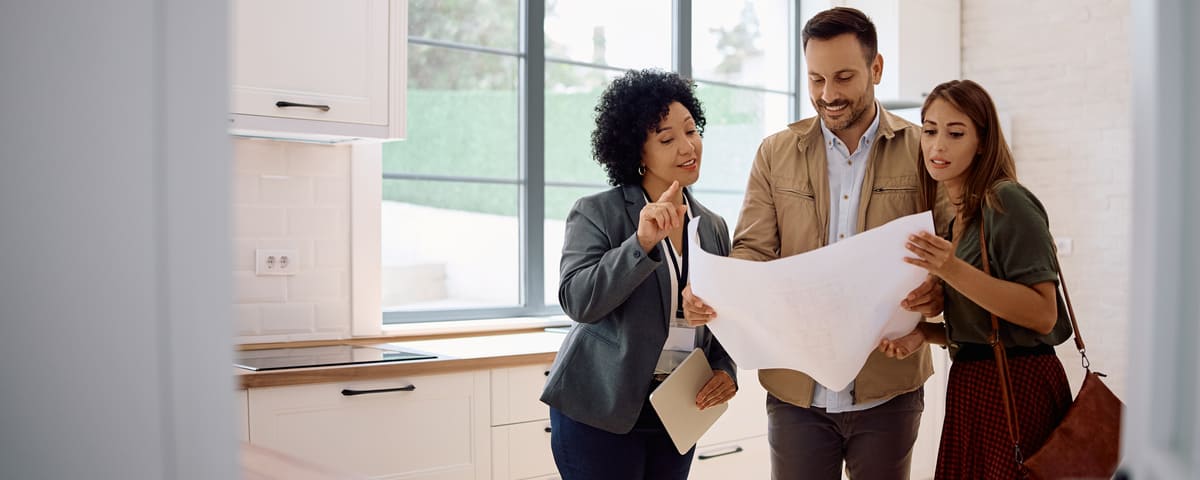 Jeune couple examinant des plans de logement avec un agent immobilier debout dans la cuisine