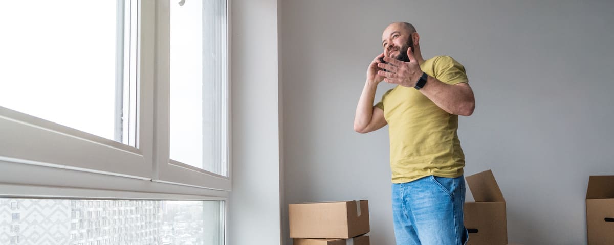 Un homme souriant debout à la fenêtre dans la pièce pleine de boîtes en carton et parlant au téléphone