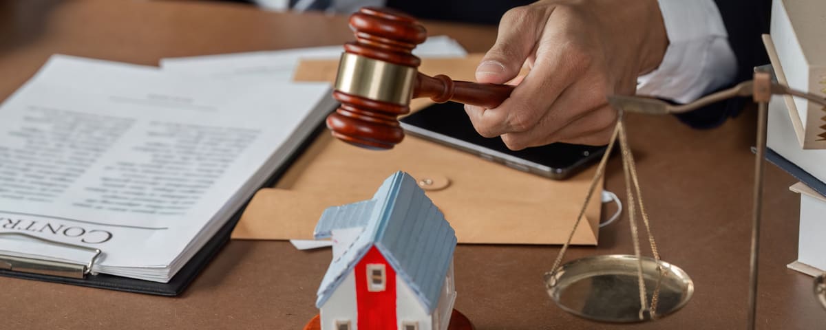 Avocat frappant le marteau sur le bureau avec une maison modèle et des échelles de justice, examinant des documents juridiques